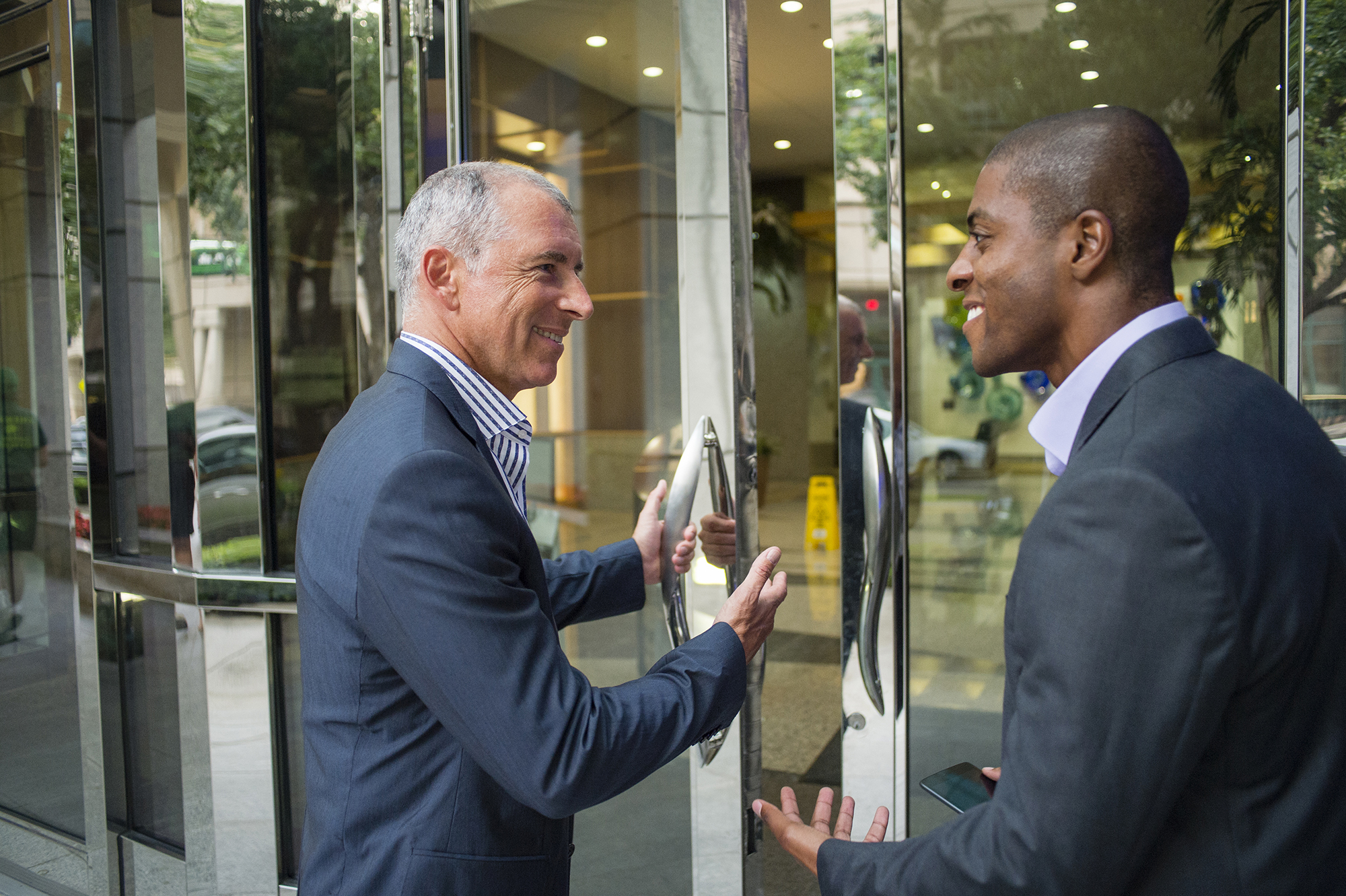 A smiling older man, dressed professionally, holds the office building door open for a fellow coworker.